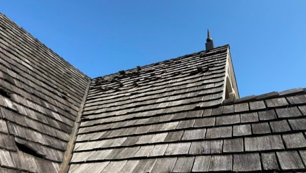 Weathered wooden shingles on a sloped roof under a clear blue sky, with some showing signs of water damage near the peak—an inviting case for water damage restoration Toronto. A chimney with a decorative cap stands in the background.