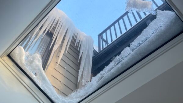 View through a skylight shows large icicles and snow buildup on the roof, with part of a deck railing outside against a blue sky—an important reminder to consider water damage restoration Toronto during harsh winters.