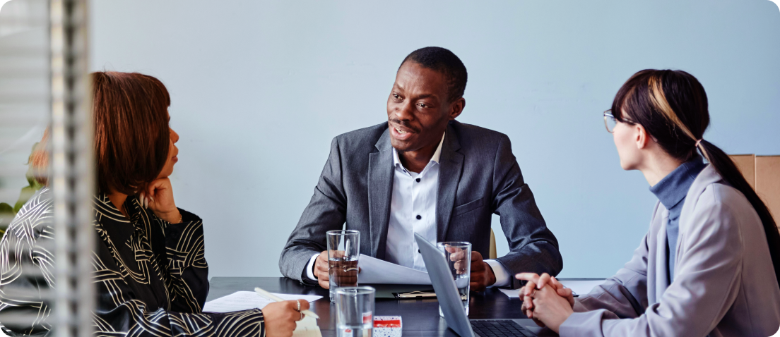 Three people in business attire sit at a table having a discussion. Two women and one man are talking, with papers, glasses of water, and a laptop in front of them. The background is plain and light-colored.