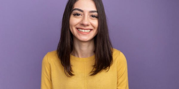 A young woman with straight brown hair, wearing a yellow sweater, smiles in front of a solid purple background.