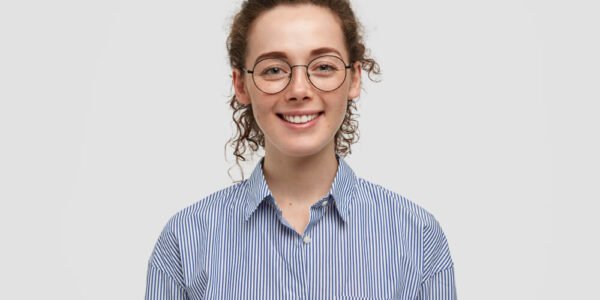 Young woman with curly hair and glasses smiles at the camera. She is wearing a blue and white striped button-up shirt and stands against a plain, light gray background.