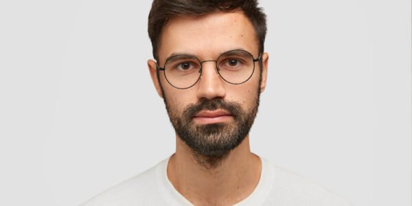 A man with dark hair, a beard, and round glasses wears a white shirt and looks directly at the camera against a plain light gray background.
