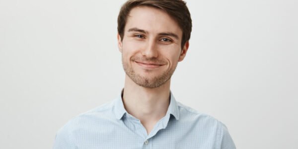 A young man with short brown hair and light stubble smiles softly, wearing a light blue checkered shirt, standing against a plain white background.