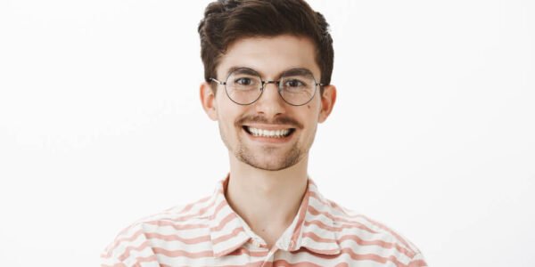 Young man with short brown hair and glasses smiles at the camera. He is wearing a light pink and white striped button-up shirt, standing against a plain white background.
