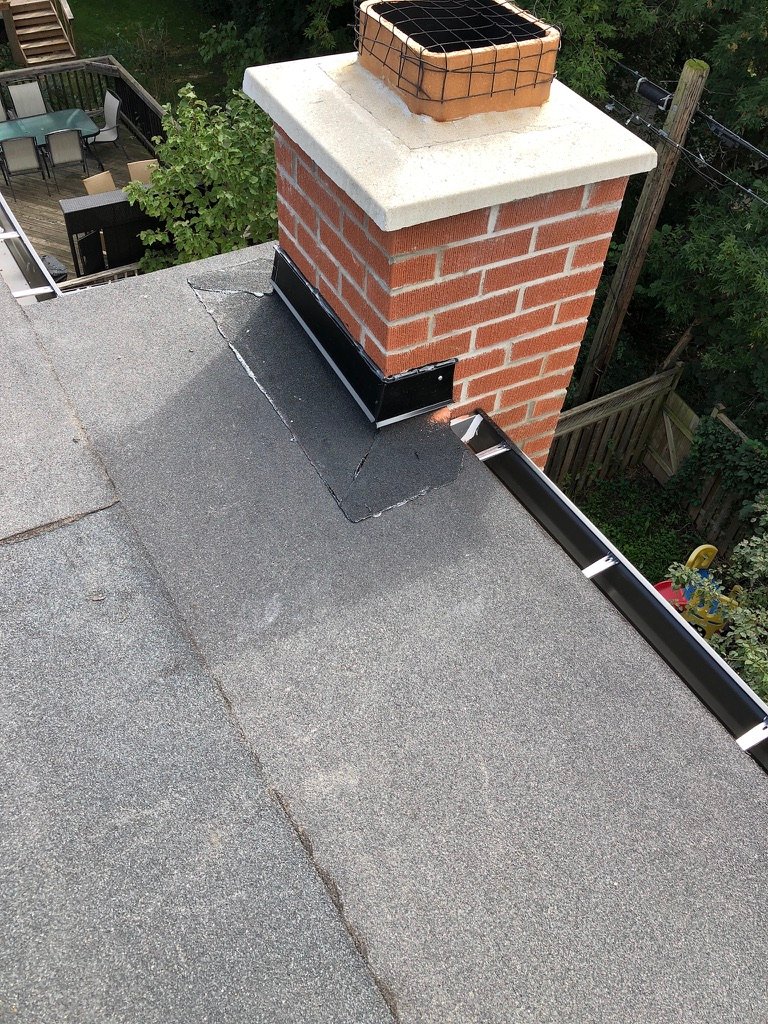 A brick chimney with black metal flashing is installed on a gray shingled roof, highlighting the quality of recent roof repair. Trees, a wooden fence, and patio furniture are visible in the background.