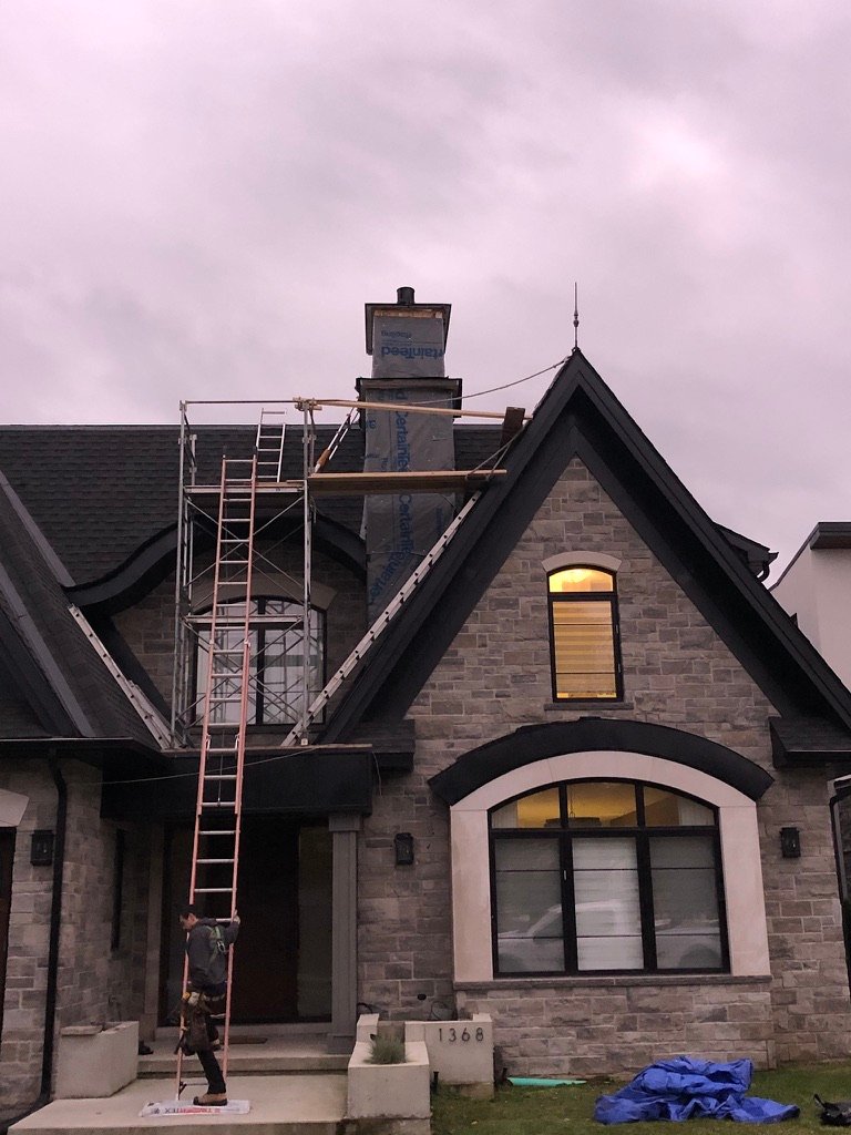 A person stands near a stone house with scaffolding and a ladder leading up to the chimney, suggesting ongoing roof repair. The sky is overcast, and several windows on the house are lit from within.