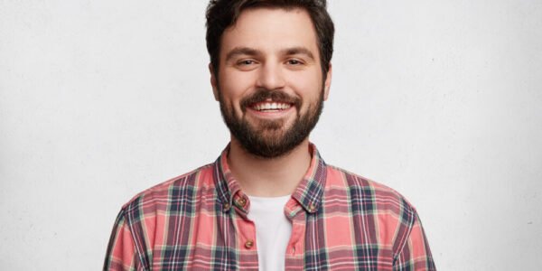 A young man with dark hair and a beard smiles at the camera, wearing a pink plaid shirt over a white t-shirt, standing against a plain white background.