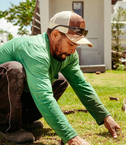 A man wearing a green shirt, cap, and sunglasses kneels on grass, carefully laying down sod patches in a yard on a sunny day.