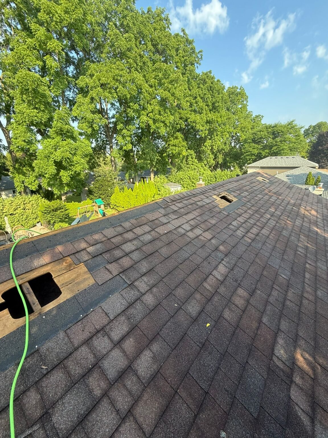 A house roof with missing shingles and two open roof vents; green hose lies across the roof. Trees and houses are visible in the sunny background under a blue sky with clouds.