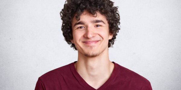 Young man with curly brown hair wearing a maroon V-neck shirt smiles at the camera, standing against a plain light gray background.