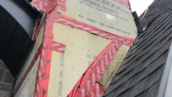 Close-up of a house exterior showing cement boards labeled USG Durock installed on a chimney with red tape sealing the seams, next to black roof shingles and a stone wall—an essential step in professional roof repair.