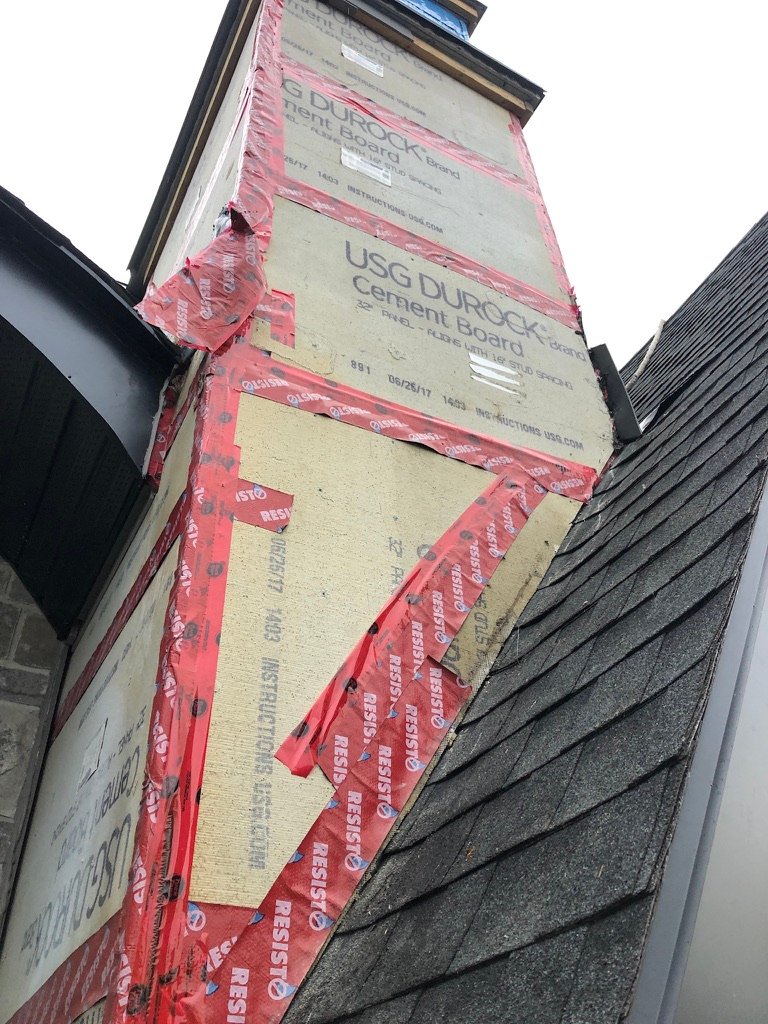 Close-up of a house exterior showing cement boards labeled USG Durock installed on a chimney with red tape sealing the seams, next to black roof shingles and a stone wall—an essential step in professional roof repair.