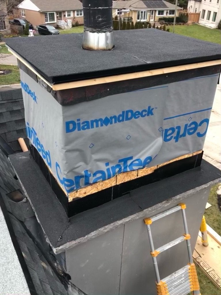 A partially finished chimney wrapped in DiamondDeck and CertainTeed underlayment, with exposed wood, black roofing material, and a yellow ladder leaning against the roof indicates ongoing roof repair. Houses and a street are visible in the background.
