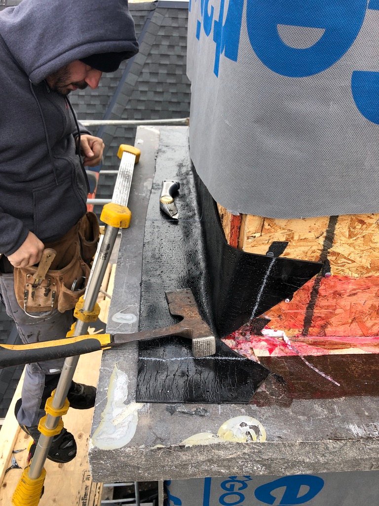A person in a hoodie and tool belt works on flashing and waterproofing around a chimney on a roof, focused on precise roof repair. Tools and construction materials are visible, and the area appears to be under renovation.