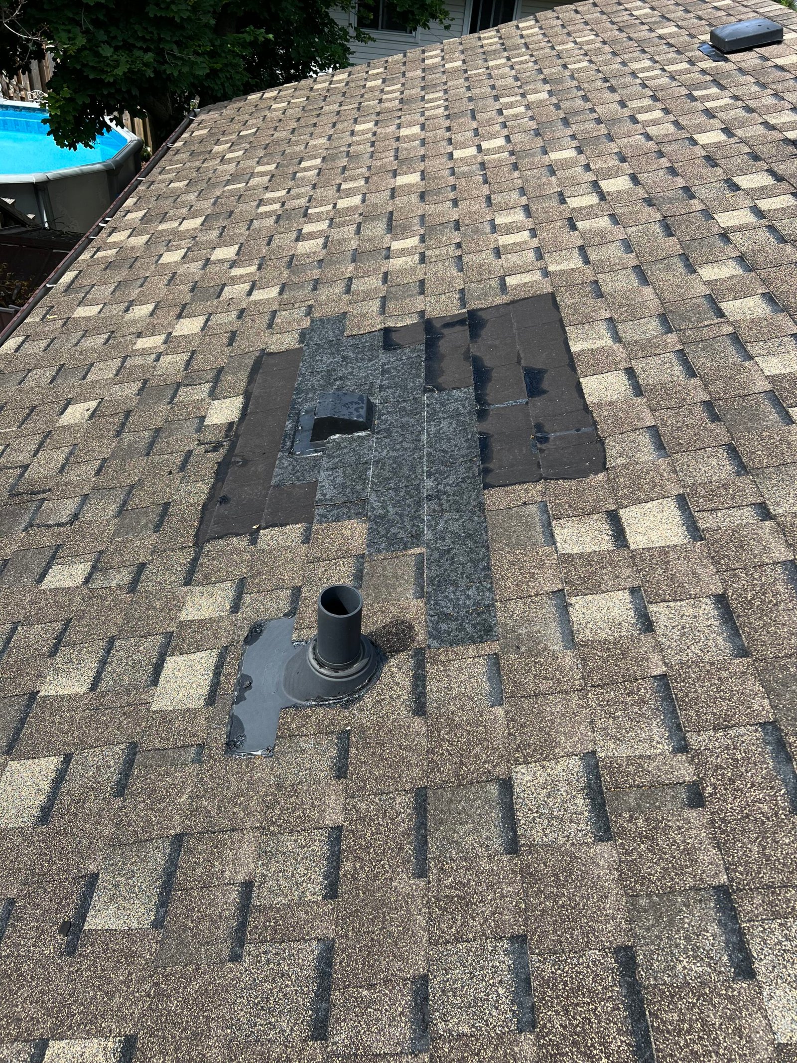 A roof with mismatched shingles showing a patch repair around a vent pipe, with different colors and patterns compared to the surrounding original shingles.
