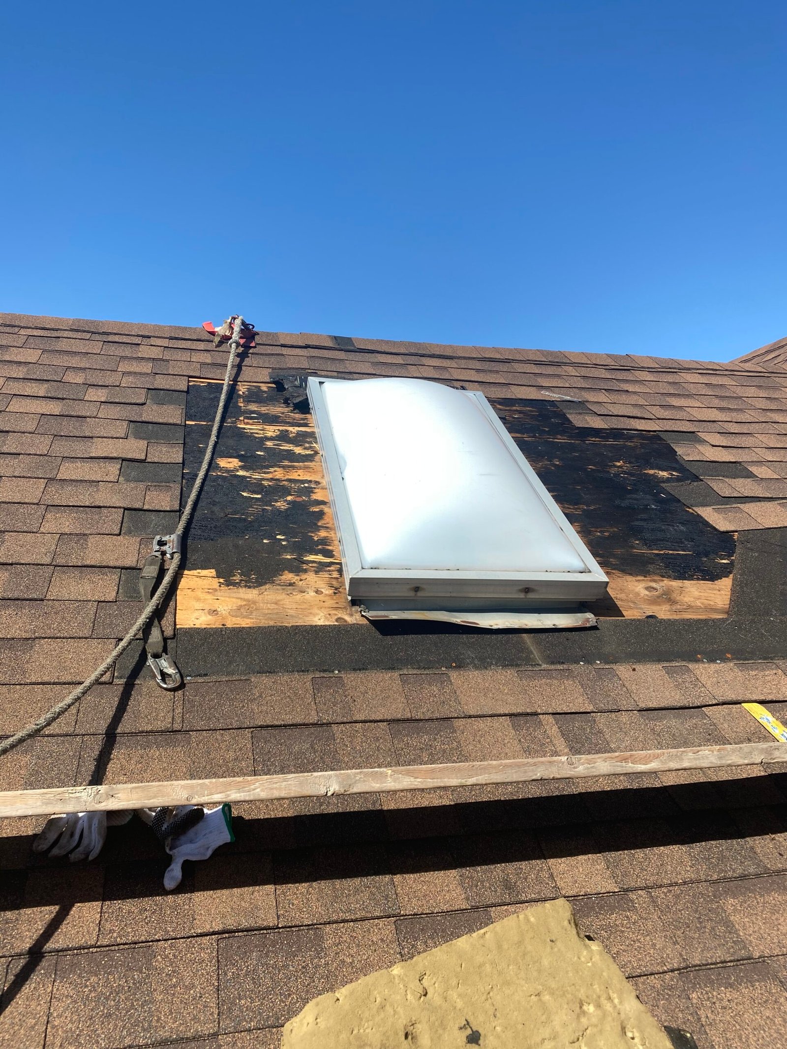 A skylight is installed on a roof with surrounding shingles partially removed, exposing black underlayment and wood. A safety rope and plank are visible on the roof. The sky is clear and blue.
