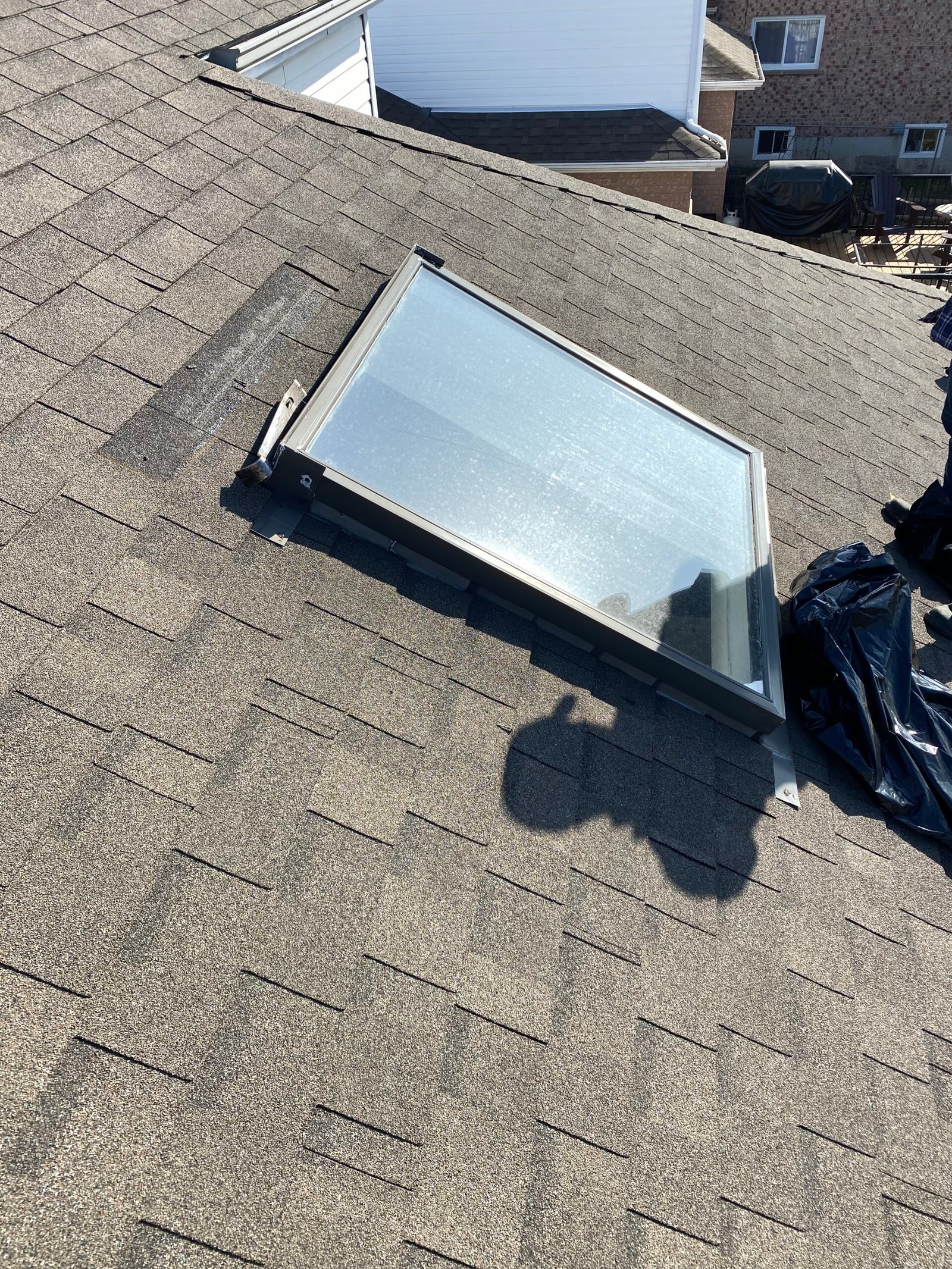 A skylight window installed on a shingled roof, with sunlight reflecting off the glass and a persons shadow visible nearby.