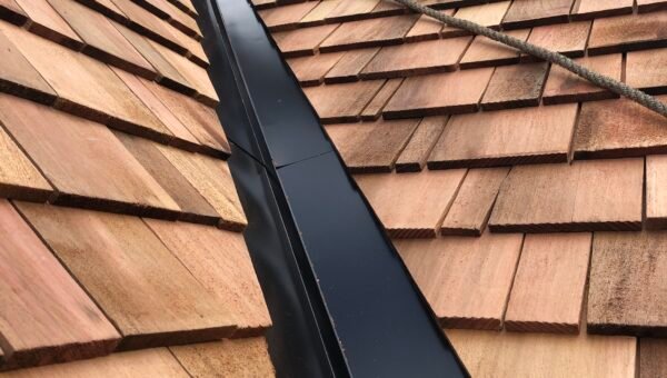Workers on a steep roof carry out roof repair as they install wooden shingles next to a black metal valley, with ropes for safety and tools visible near the top of the roof.