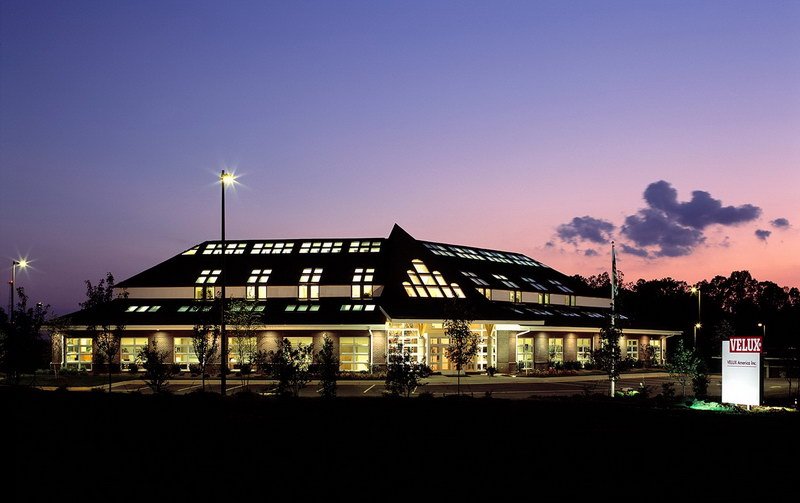 A modern, glass-paneled building is lit up at dusk with a purple sky in the background. The sign in front of the building reads VELUX. Streetlights and trees surround the building.