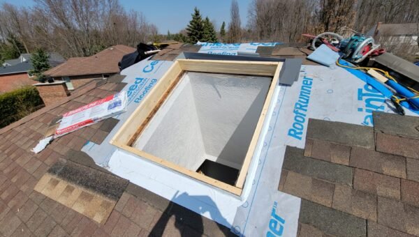 A roof with a rectangular skylight opening surrounded by roofing underlayment, roofing tools, and shingles, with trees and neighboring houses visible in the background under a clear sky.