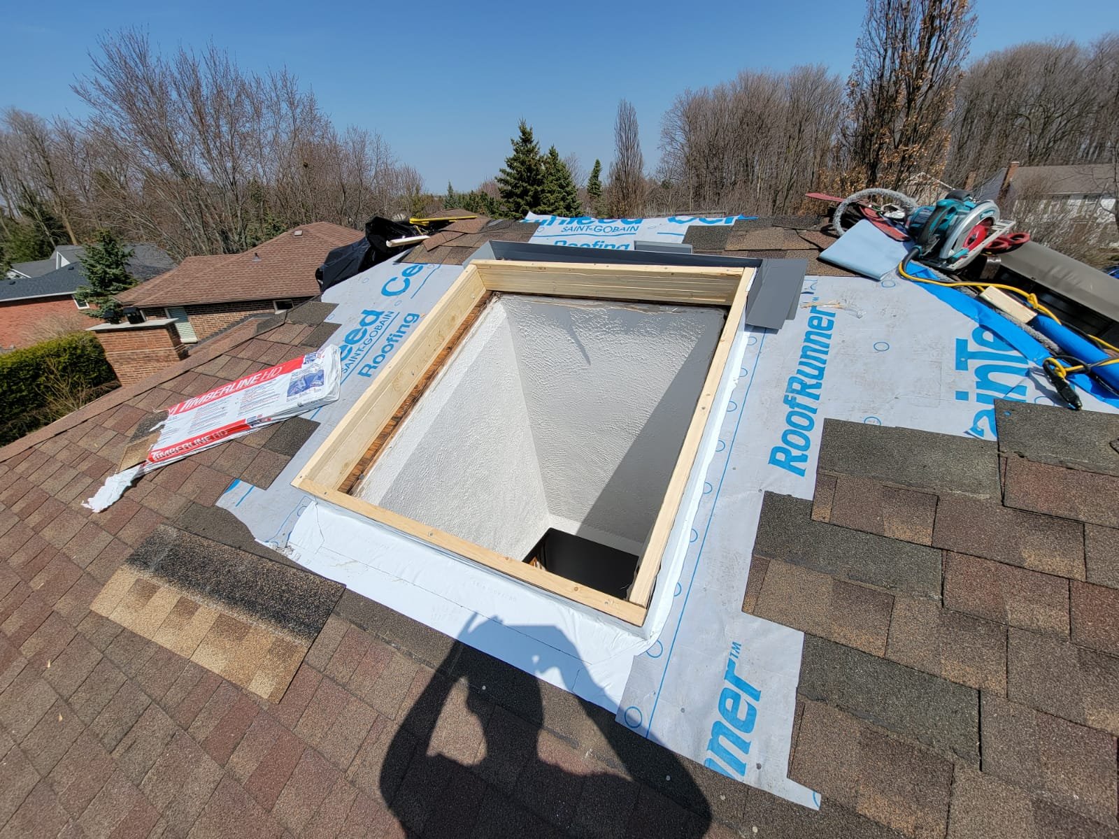 A roof with a rectangular skylight opening surrounded by roofing underlayment, roofing tools, and shingles, with trees and neighboring houses visible in the background under a clear sky.