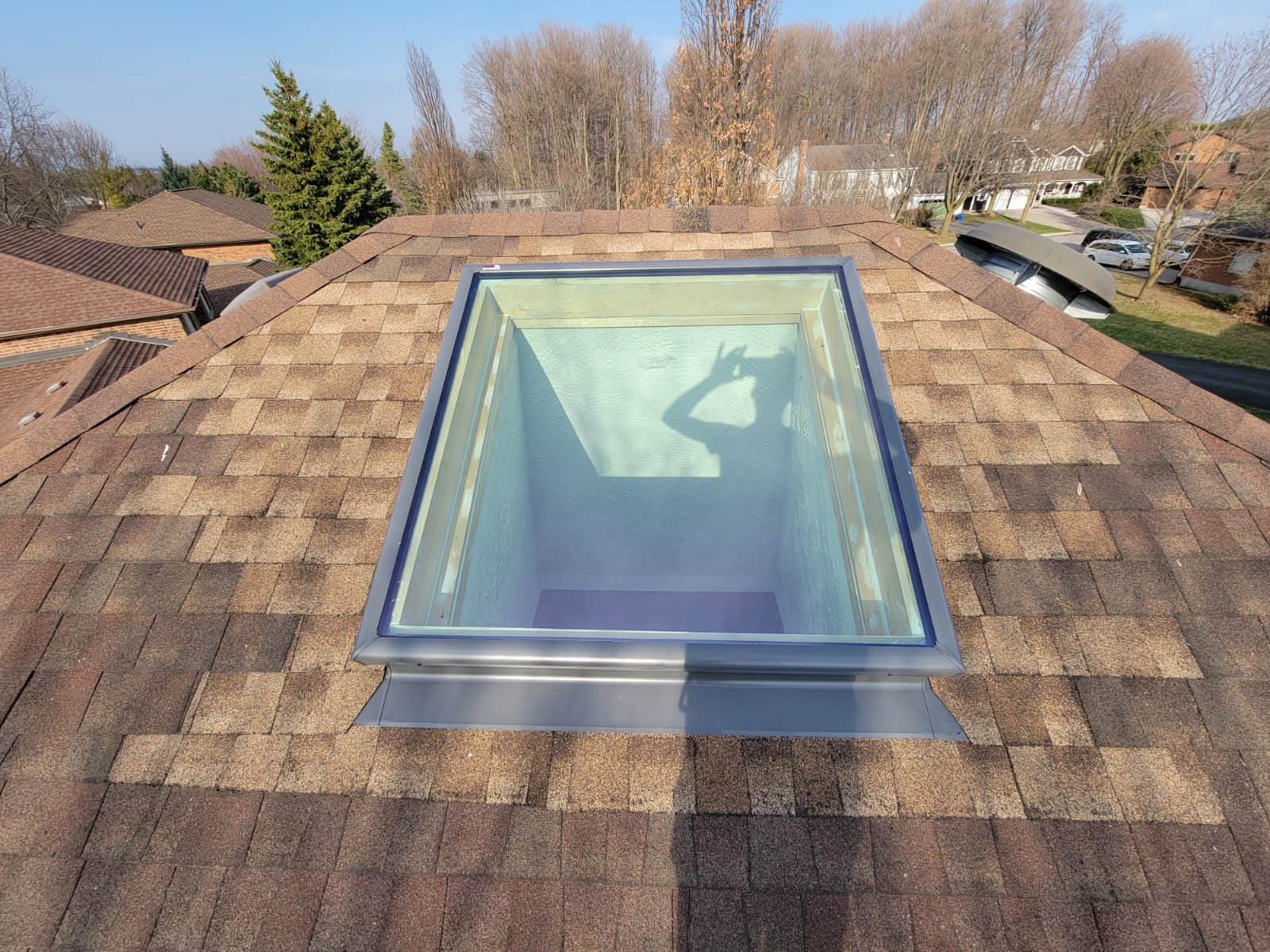 A skylight window installed on a brown shingled roof, reflecting a person making a hand gesture shaped like an OK sign, with trees and houses visible in the background.