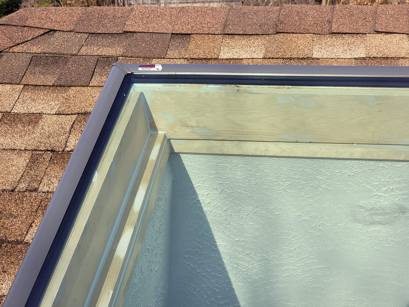 Close-up of a skylight frame installed on a roof with brown shingles, showing wood framing, glass panel, and textured white interior surface beneath the skylight. Trees are visible in the blurred background.