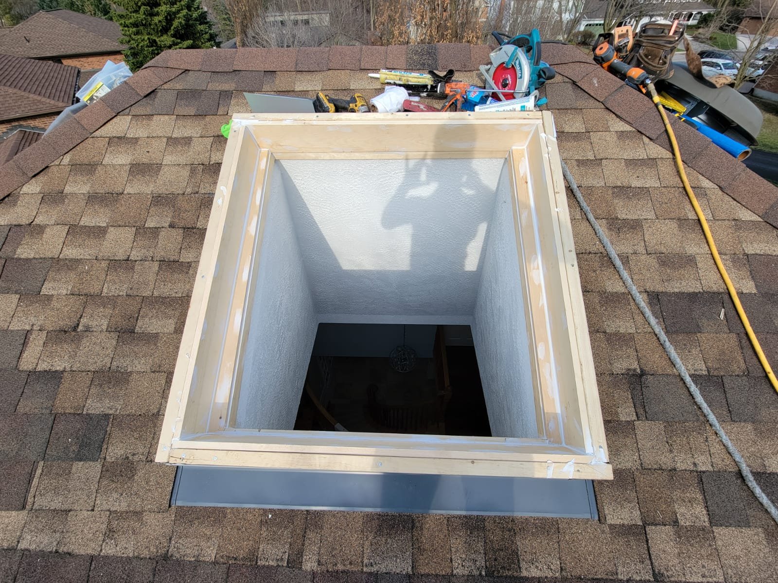 A rectangular skylight opening on a shingled roof, surrounded by wood framing, with tools and safety rope nearby. A persons shadow is visible at the edge of the opening.
