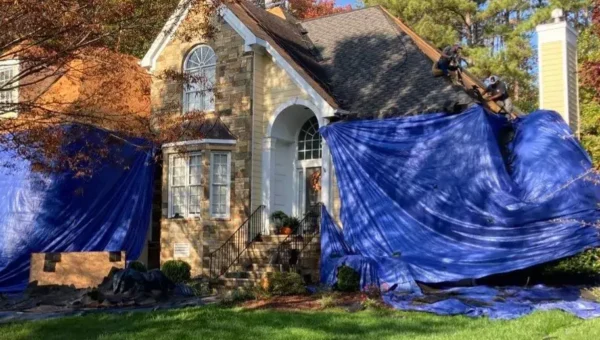 A house with part of its exterior and roof covered in large blue tarps. Workers in safety gear are on the roof, possibly performing repairs. The yard is green and leafy trees are nearby.