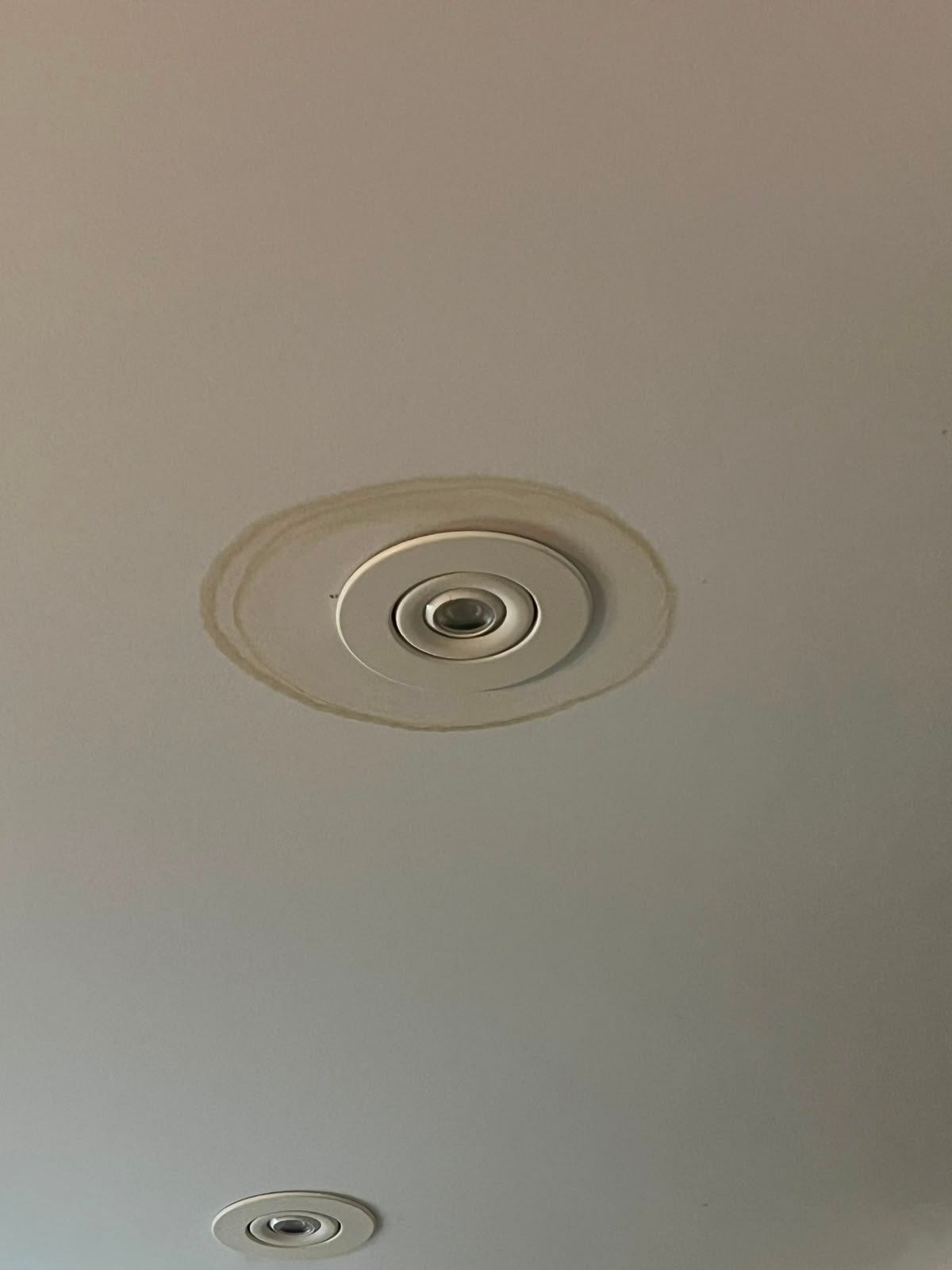 A recessed ceiling light surrounded by a brown water stain, indicating possible water damage; another light is partially visible in the lower part of the image.
