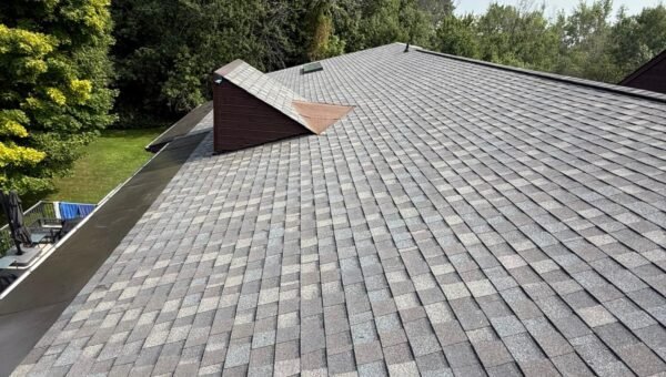 A view of a sloped roof with gray asphalt shingles, a small dormer, and trees in the background under a partly cloudy sky.