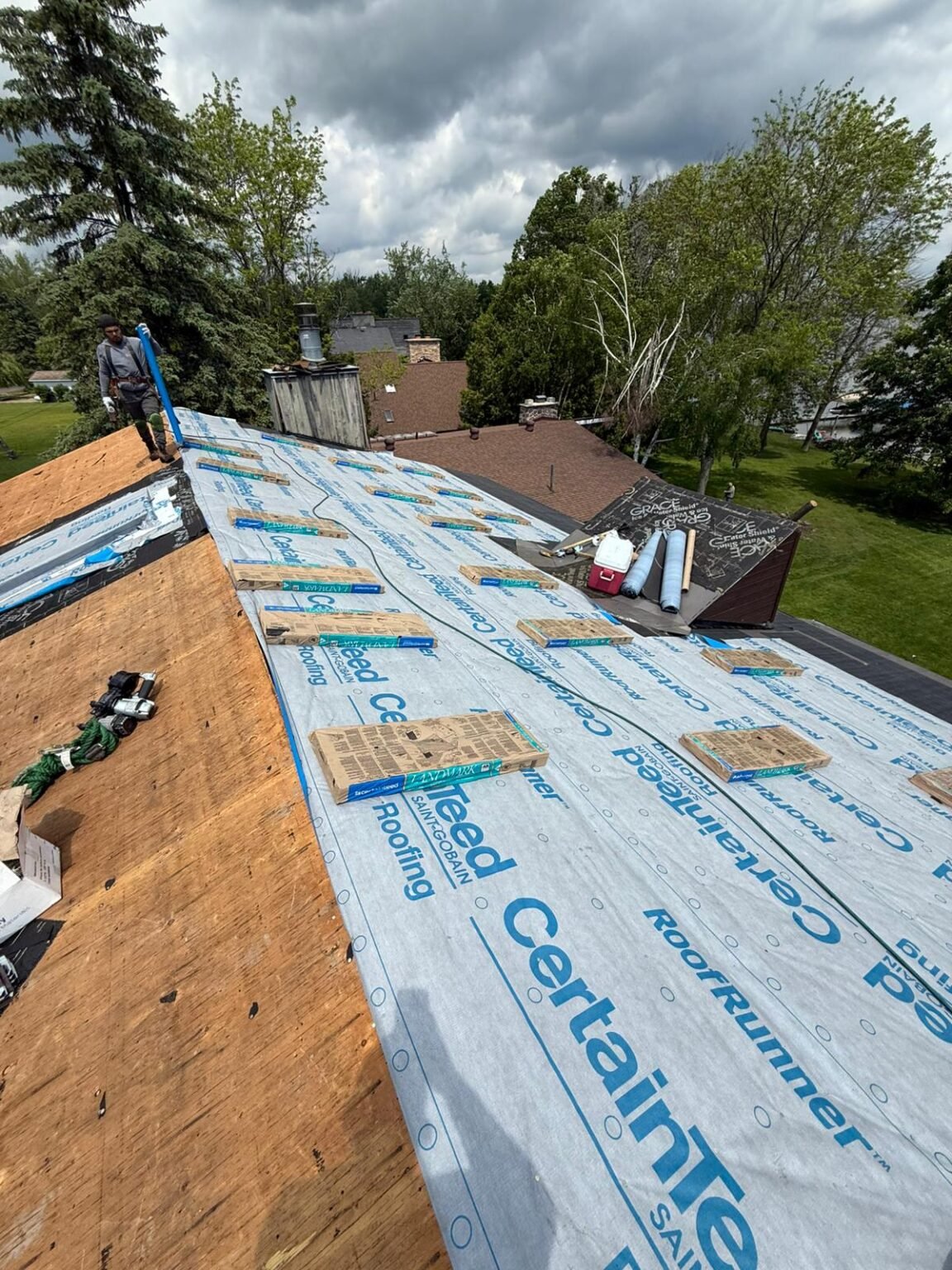 A person works on a partially covered house roof, installing CertainTeed underlayment. Wooden boards, tools, and roofing materials are scattered around. Trees and a neighboring house are visible in the background under a cloudy sky.