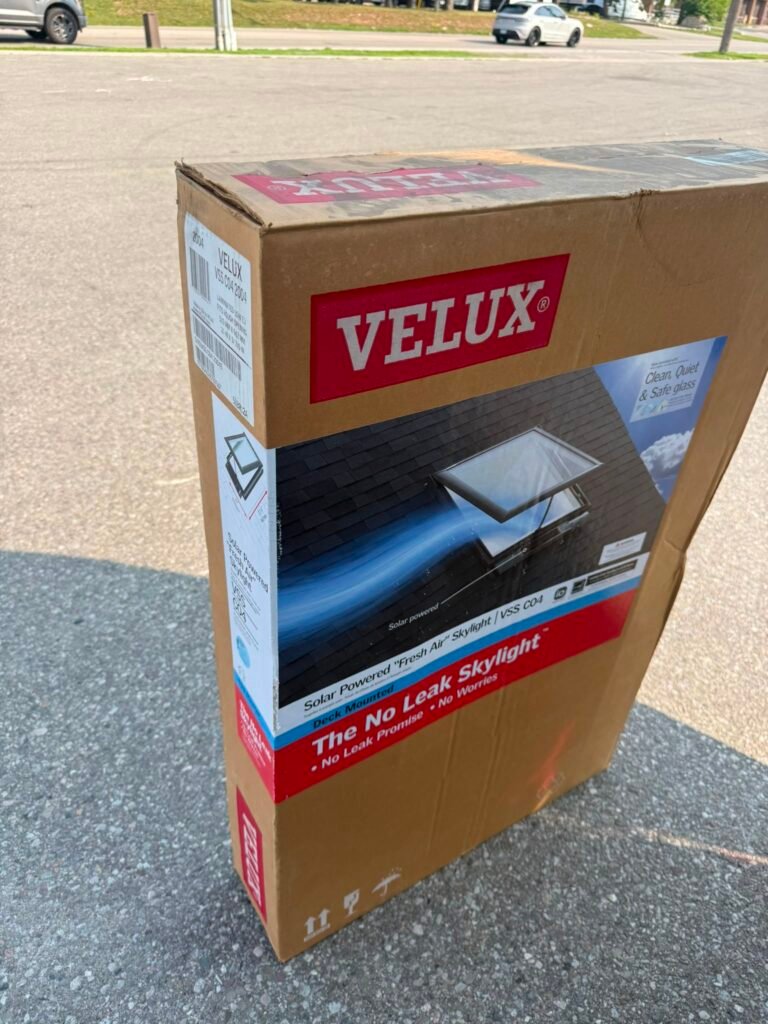 A sealed box of a VELUX solar-powered No Leak Skylight is shown standing on a sunlit pavement, with product details and an image of the skylight on the packaging.