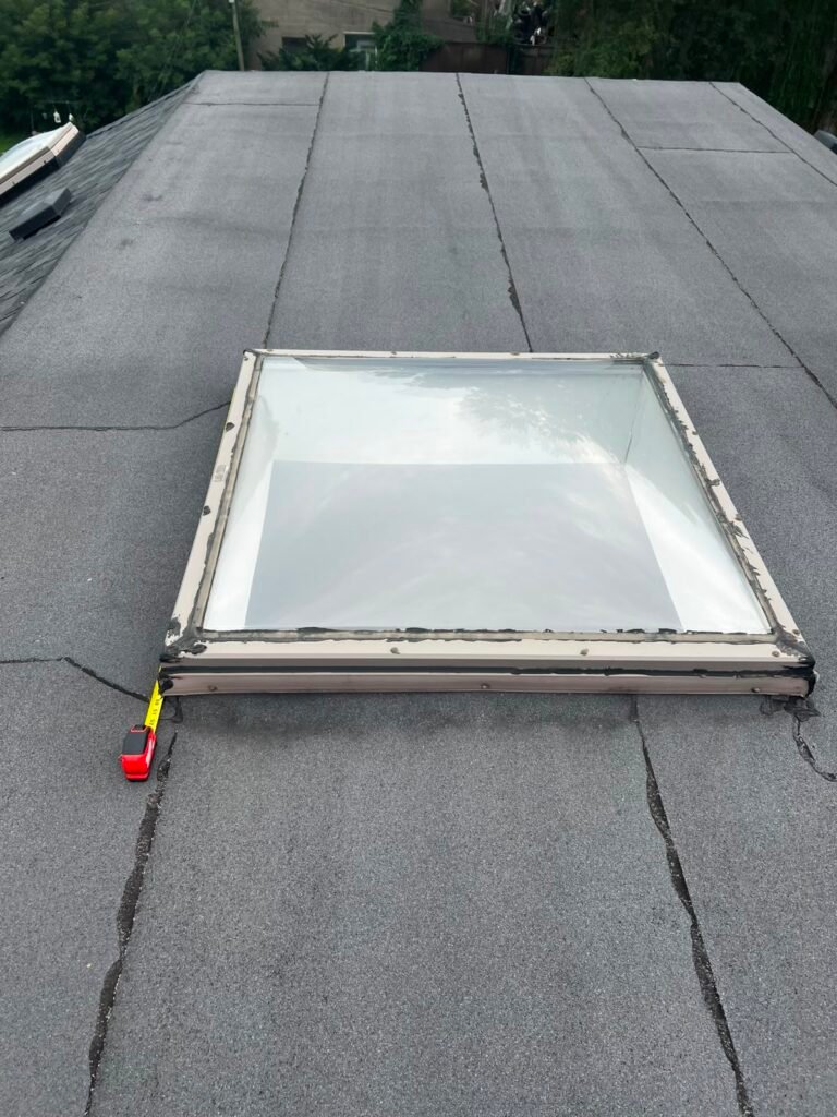 A square skylight sits on a flat, gray, asphalt roof. A red and yellow tape measure is beside the skylight. There are visible cracks on the roof surface around the skylight.