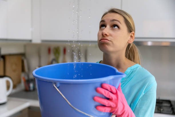 A woman wearing pink gloves holds a blue bucket under a leaking ceiling in a kitchen, looking up with a frustrated expression as water drips down.